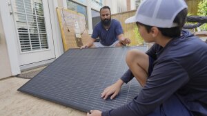 Squatting near the ground outdoors, Bhavin Misra and his son, Rumi, hold opposite sides of a black, square solar panel with a grid pattern on it while assembling a plug-in solar kit at their home in Houston.