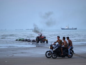 Young men ride motorcycles along the shore as a tractor pulls equipment from the water and commercial vessels appear anchored in the Strait of Hormuz off Bandar Abbas, Iran, Monday, July 27, 2026.