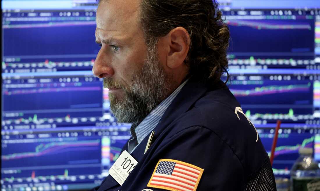 A trader works on the floor of a stock exchange. He is wearing a navy blue blazer, and screens displaying stock market information are behind him.