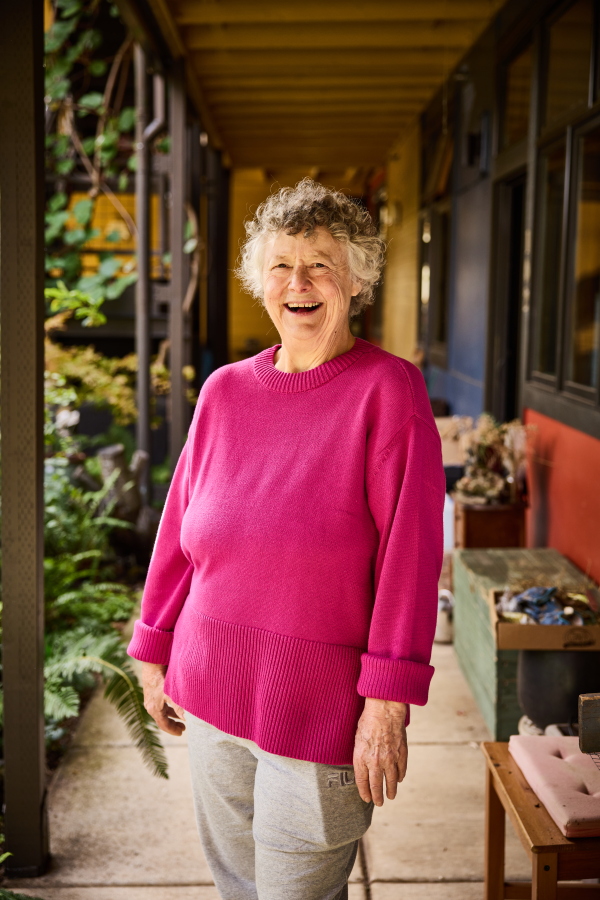 Deana Camp, 73, has curly gray hair and is wearing a bright pink sweater. She is smiling broadly.