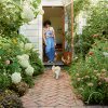 A photograph shows chef and cookbook author Samin Nosrat standing in the doorway of a house, colorful art hangs on the wall behind her, the glass door opens to her right, and she's surrounded by tall, leafy hydrangeas, allium, ferns, and other plants on either side. Samin is barefoot, wearing a colorful button-up shirt, leaning casually against the doorframe, and smiling down at a small white dog walking toward her on a brick pathway.
