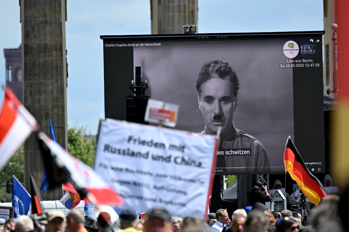 Demonstrators in Berlin wave flags and hold a poster reading 'Peace with Russia and China' as they rally in front of a screen displaying a scene from the movie The Great Dictator during the 'Peace - Freedom - Referendum' demonstration in May 2025.