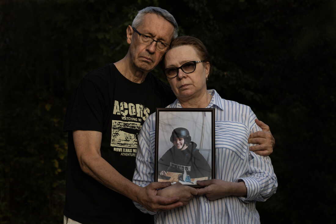Andriy Kuvshynov (left) and Iryna Mamaysur hold a framed portrait of their daughter, Oleksandra Kuvshynova, in 2024. Kuvshynova is wearing a helmet and a protective vest that says "Press." Kuvshynov has his arm around his wife.