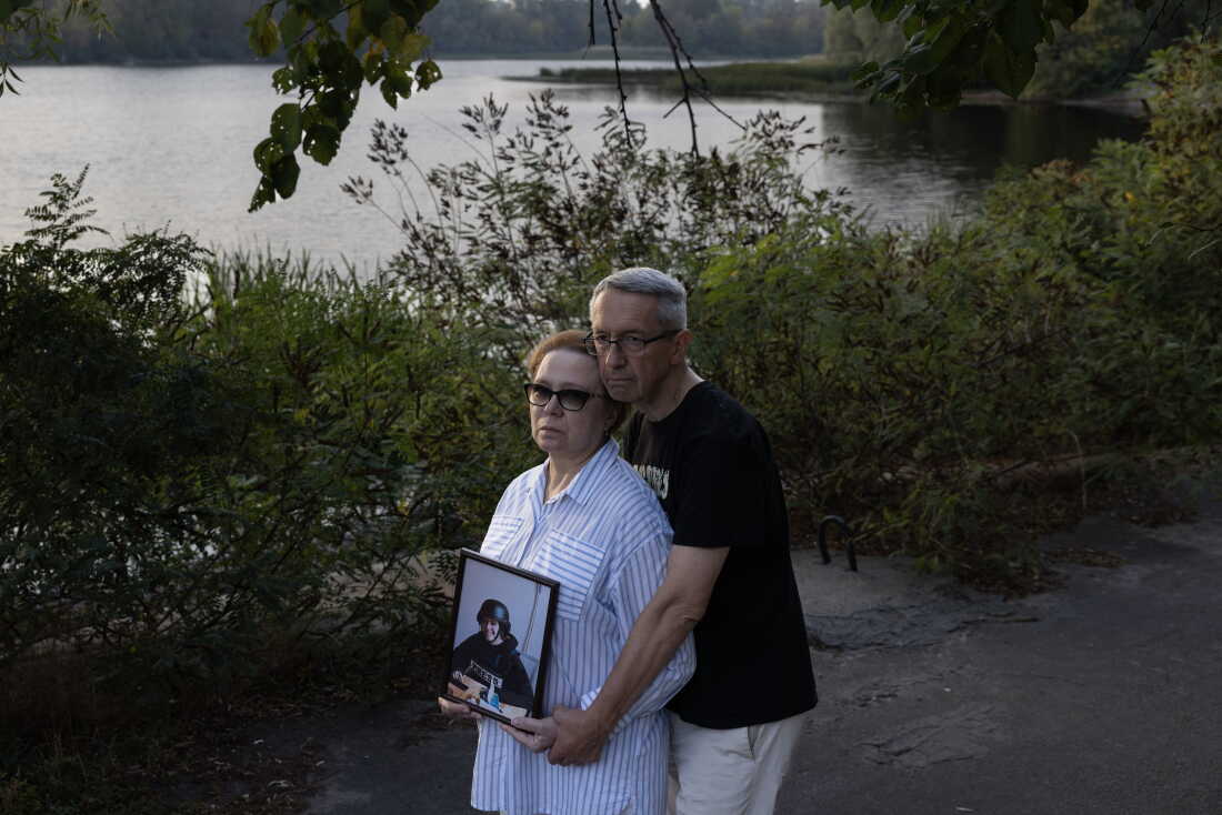 Iryna Mamaysur holds a framed photo of Oleksandra Kuvshynova. Andriy Kuvshynov is embracing her from behind. They are standing by the water in Kyiv.