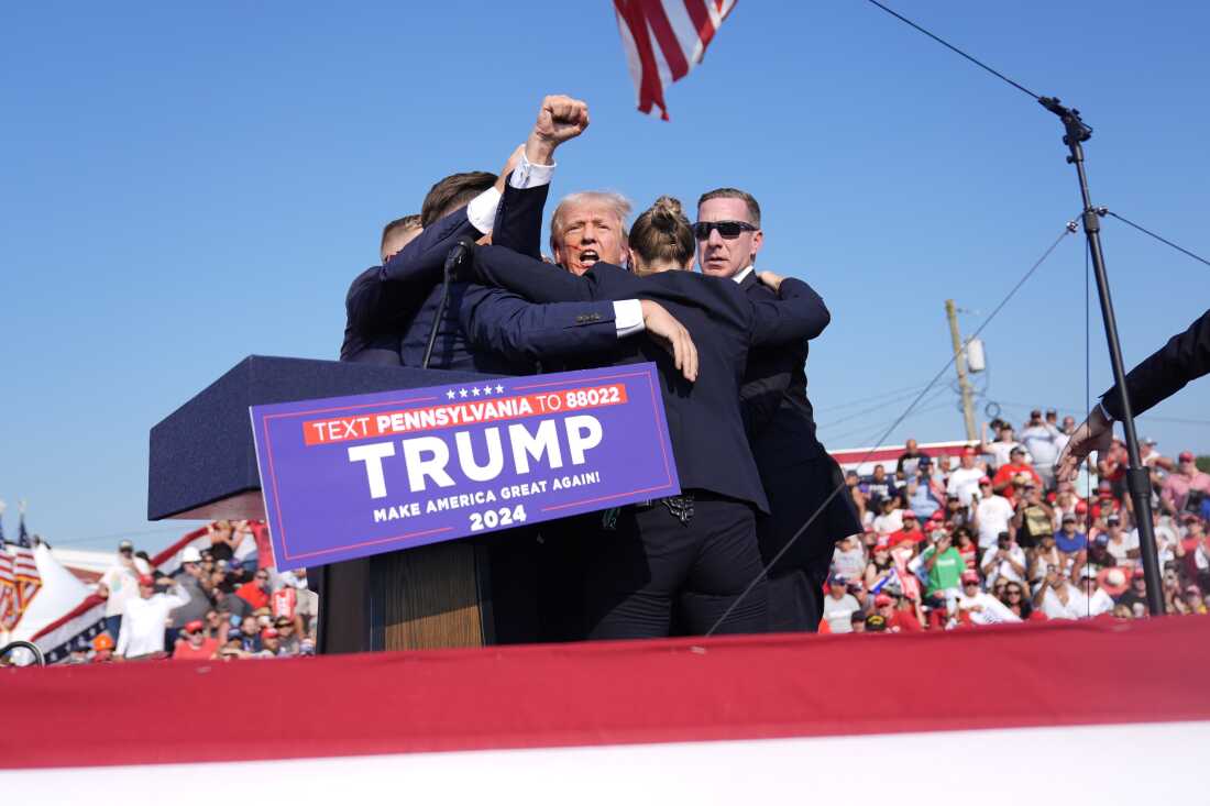 Trump is holding a fist in the air as Secret Service agents surround him as he leaves the stage at the campaign rally in Butler on July 13. The agents are wearing dark blue suits. Behind the stage are rally attendees.