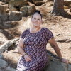 In this photo, Lisa Collenbaugh is sitting on a large rock outdoors. She's wearing a long dress with short sleeves that's black with a pink and tan pattern on it.