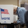 A man leans over to fill out a ballot inside a voting booth that says "Vote" on the side with a U.S. flag.