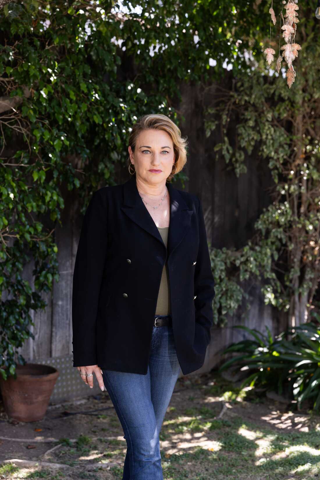 This photo is a portrait of Naomi Seligman. She's standing outdoors, with a fence and green foliage behind her. She's wearing blue jeans, an olive shirt and a black blazer.
