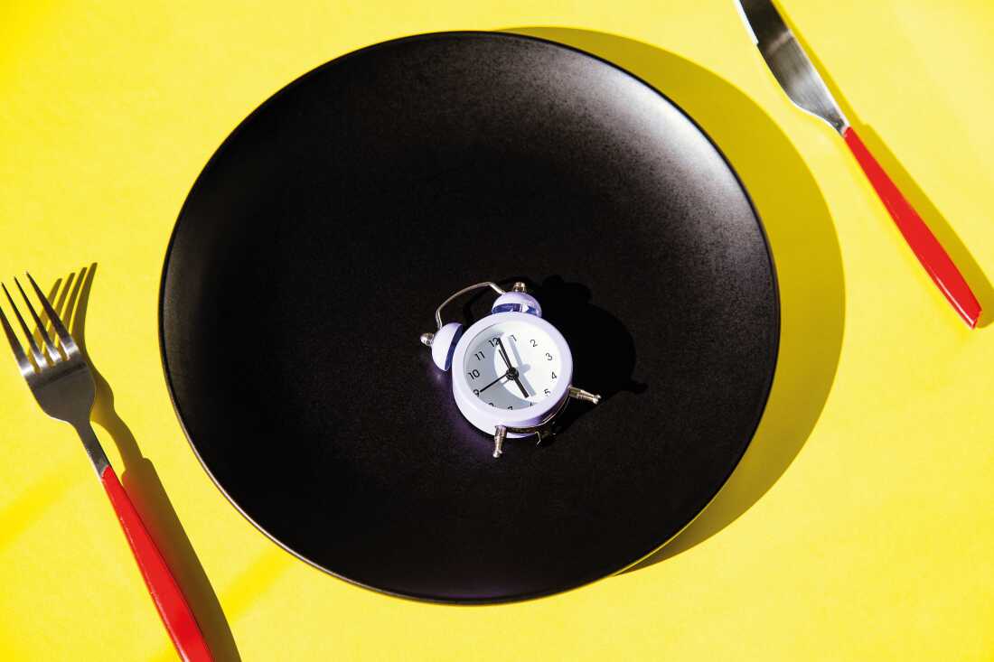 Studio photograph shows a large, black, ceramic dinner plate on a yellow background. A metal fork and knife with red plastic handles are on the right and left of the plate and a a small, white alarm clock rests on the black plate.