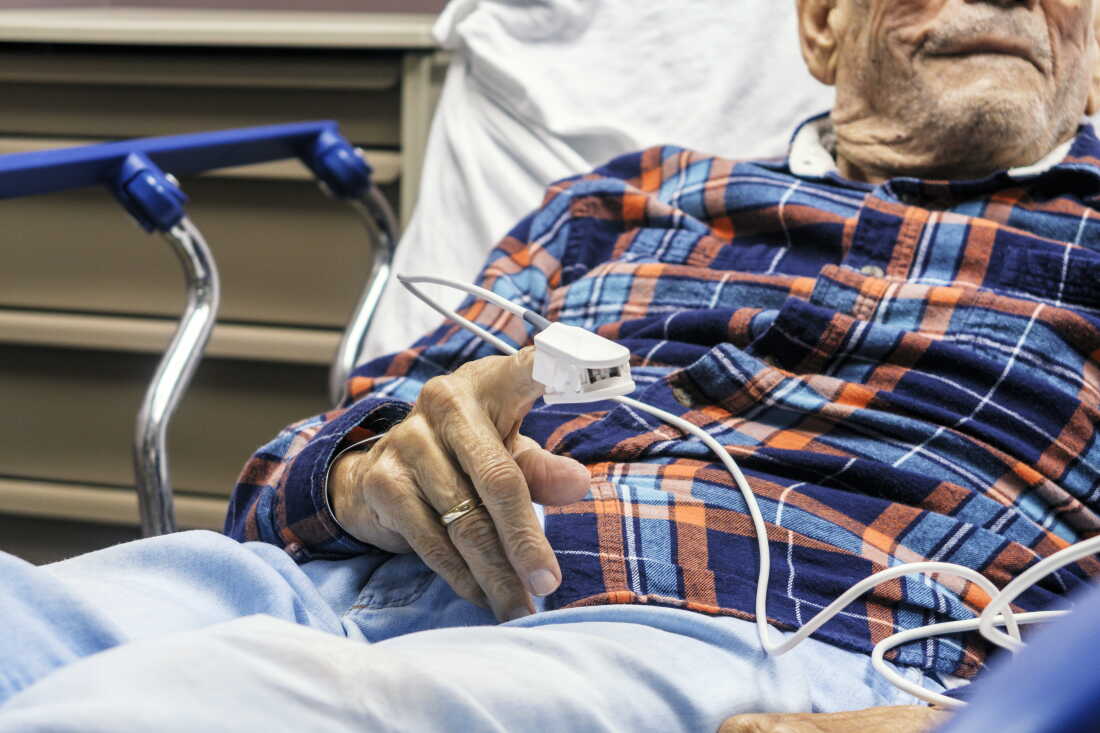 An older man lies in a hospital bed with guardrails. He is wearing a blue and red plaid shirt and having his pulse oxygen level measured with a device on his finger. His face is only partially visible.