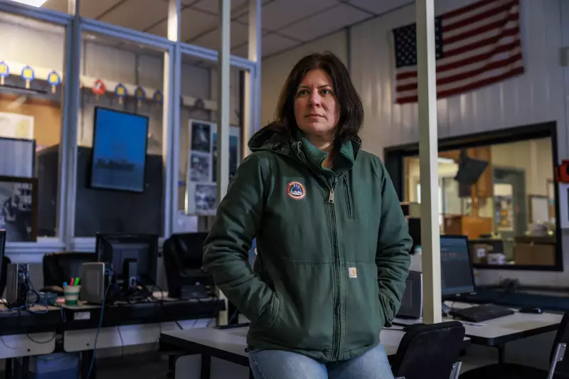 Wearing a green jacket and blue jeans, Mary Hudson is sitting on a desk inside a warehouse. Around her are other desks and workstations that have computer monitors on them.