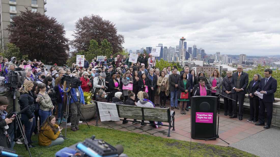 People are shown gathered around a podium where a person is speaking. The skyline of Seattle is visible beyond them.