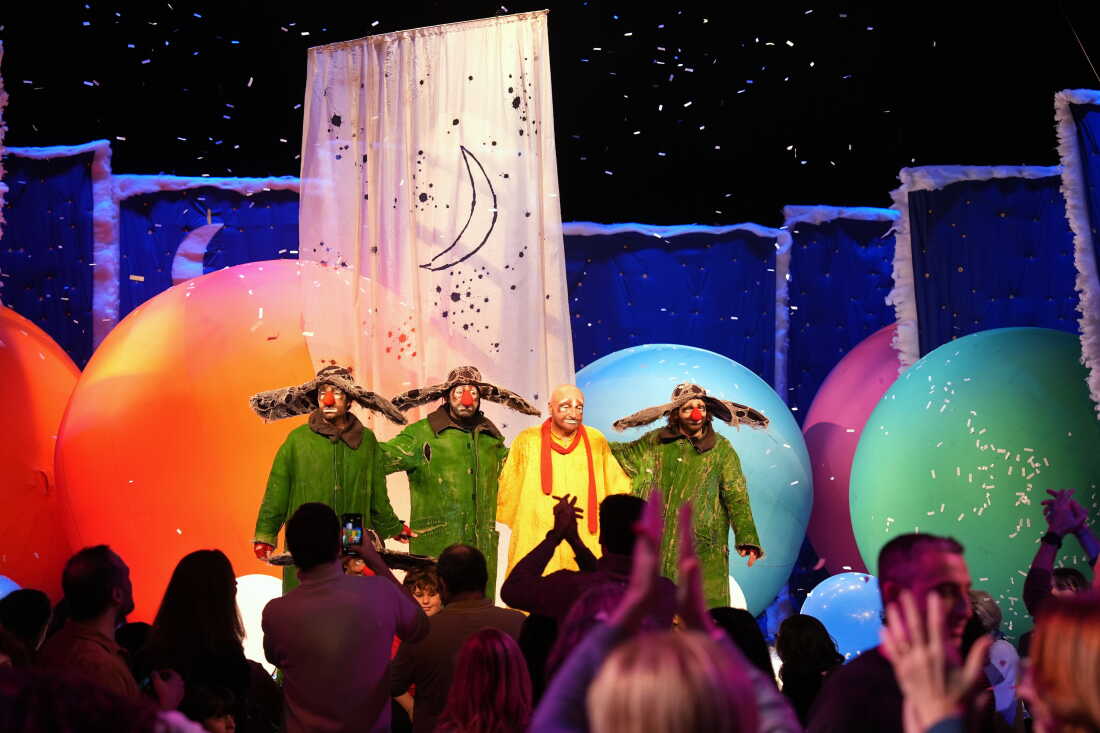 Slava Polunin performs on a stage with three members of his company. All four are wearing red clown noses; three performers are wearing green costumes, and one is wearing a yellow costume. Giant multicolored balls are behind them.