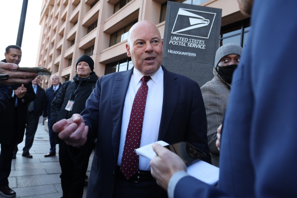 In this 2022 photo, Louis DeJoy, then the U.S. postmaster general, speaks to reporters. He's wearing a dark blue suit and is standing outside the U.S. Postal Service headquarters building while surrounded by reporters.
