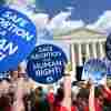 The image shows a bright blue sky and fluffy clouds above the Supreme Court building in the background, and protestors holding blue signs with white type that read, "Safe abortion is a human right" and "Keep abortion legal" in the foreground.