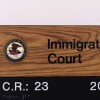 This file photo shows a rectangular sign that indicates the location of an immigration court in New York City. The sign says "Immigration Court" on a wood-patterned background, and the round Department of Justice seal, with an eagle on it, is toward the top-left corner. The bottom of the sign says "C.R.: 23" and "2035," with braille underneath.
