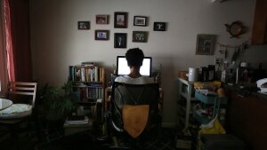 A woman is shown from behind at a sitting at a desk facing a computer screen. An array of small, framed photographs are on the wall above the computer.