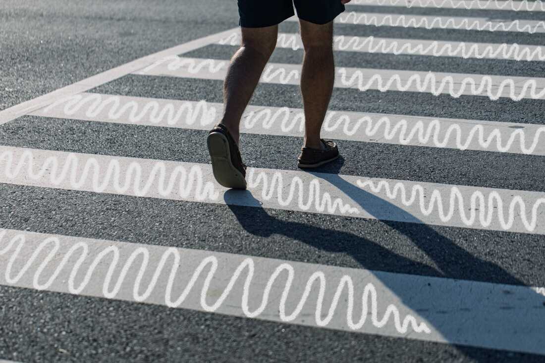 A close-up photograph shows nan little half of a personification wearing shorts and vessel shoes stepping crossed a achromatic striped crosswalk connected an asphalt road. A beardown protector is formed to nan correct of nan personification and achromatic zig zag doodles stress nan crosswalk stripes. 