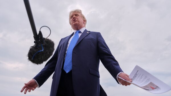 President Trump speaks to reporters who are not seen in the image. He wears a dark blue suit and a lighter blue tie, and holds a set of papers in one hand.