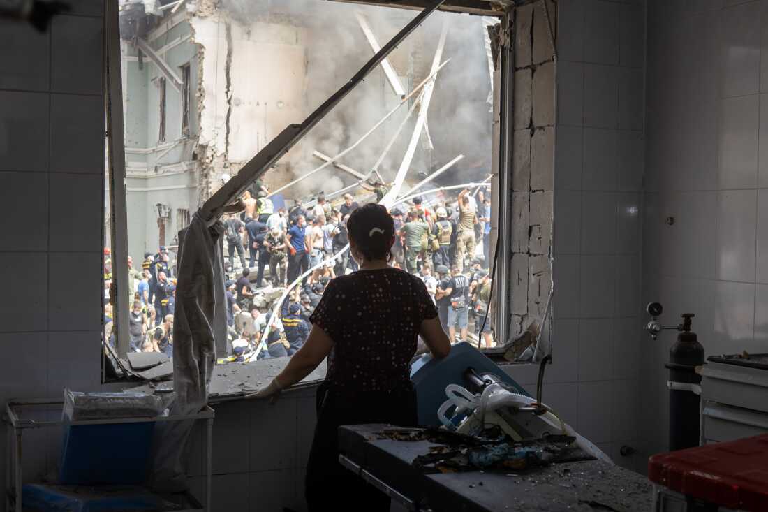 A health care worker looks through a window of a damaged operating room as people clear rubble at the building of one of Ukraine's largest children’s hospitals, Okhmatdyt, partially destroyed by a Russian missile strike on Monday.