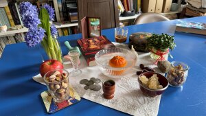 Objects including purple hyacinthsa mirrora bookcoins and an orange floating in a glass bowl sit atop a white cloth with silver embroidery.