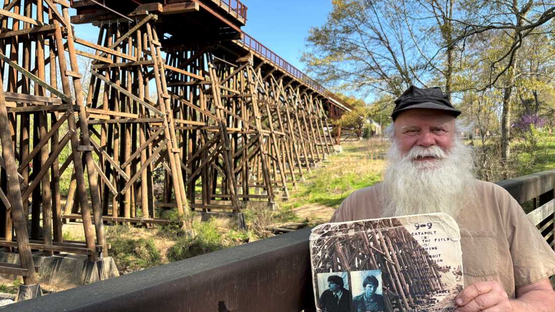 Paul Butchart memberikan tur landmark musikal di seluruh Athena, Ga. Di tangannya adalah album murmur Rem 1983, dengan foto Trestle Bridge asli di sampul belakang. Jembatan di belakang Butchart adalah rekonstruksi, dan sekarang bagian dari jalan setapak dan bersepeda.