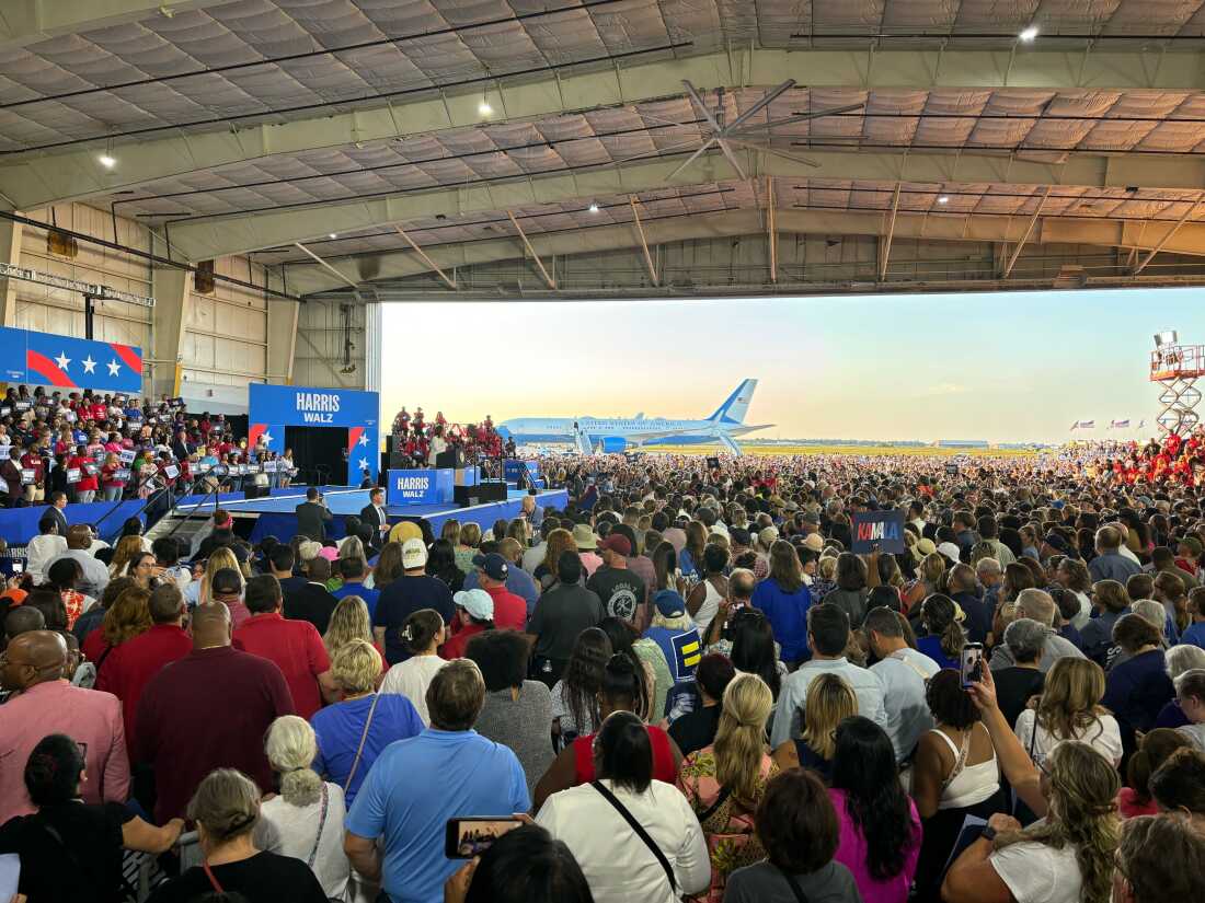 This photo shows a scene from a Kamala Harris and Tim Walz rally in Detroit on Aug. 7. Many people are crowded together in an aircraft hangar, facing a stage where Harris speaks at a lectern. The hangar is open to the outdoors, and outside is Air Force Two and a blue sky.