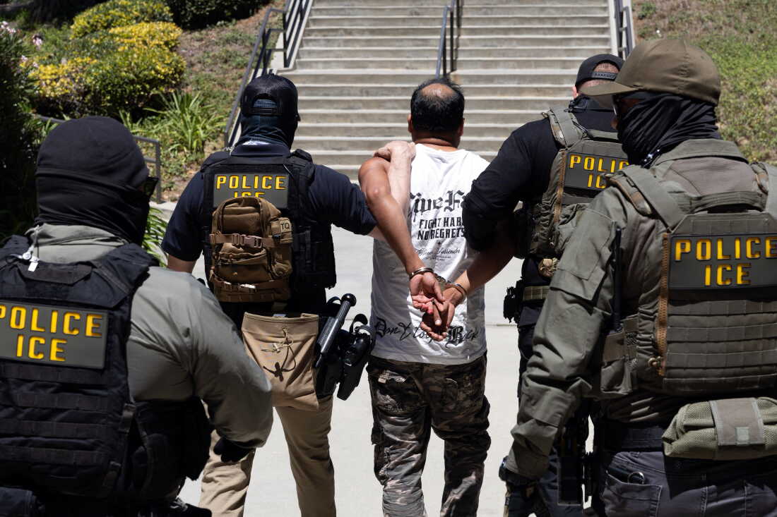 Four Immigration and Customs Enforcement agents walk a detainee out of the Ventura County Government Center in Ventura, California, on July 27. The individuals are photographed with their backs to the camera, and the detainee is handcuffed behind his back. The agents are wearing bulletproof vests that say "POLICE ICE" in yellow capital letters.