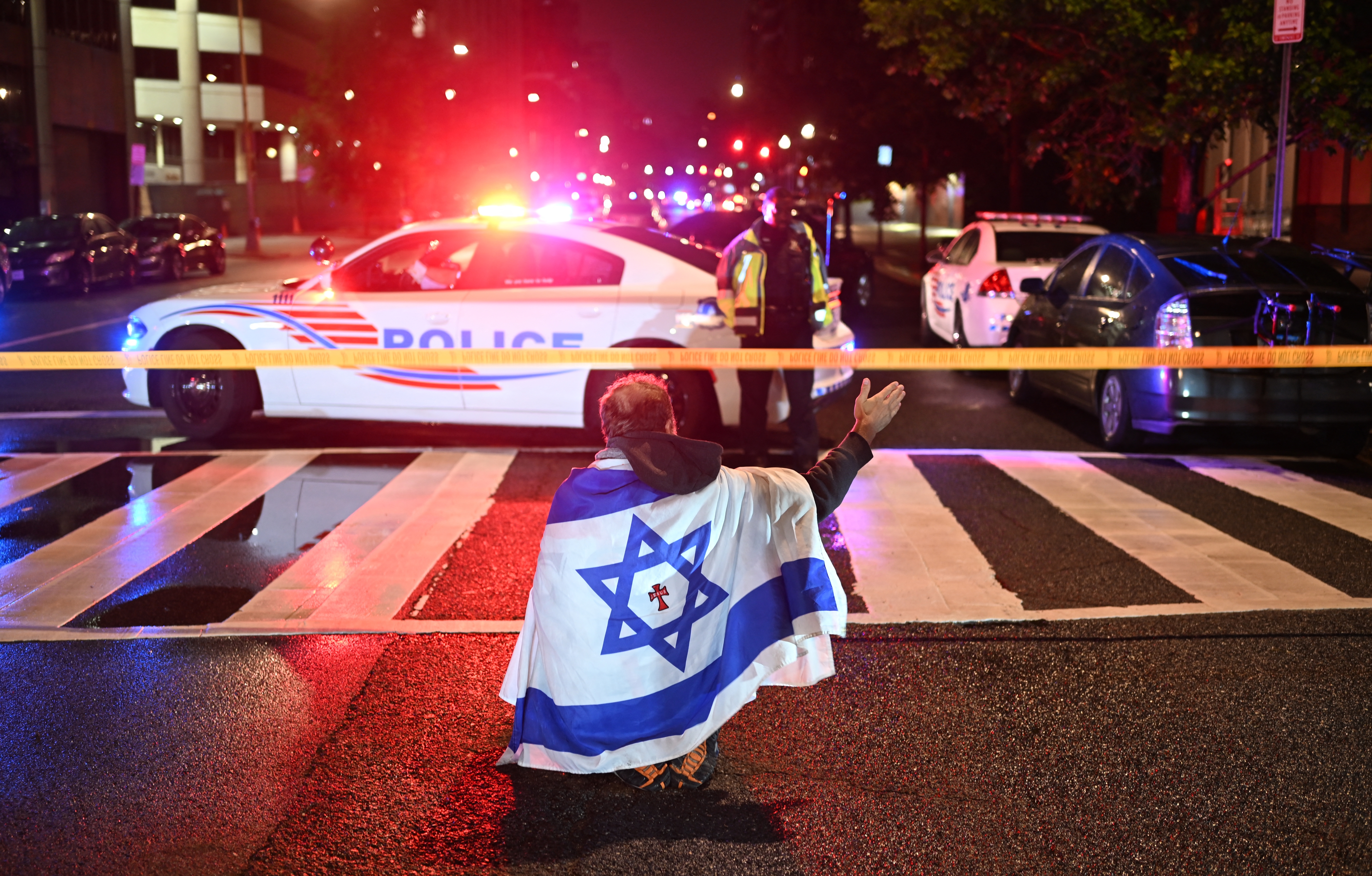 A man draped in the Israeli flag, bearing a cross and the name "Jesus" at its center, gestures as Metropolitan Police officers secure the area outside the Capital Jewish Museum in Washington, D.C. Two Israeli embassy staffers were shot dead late Wednesday by a gunman who allegedly shouted "free Palestine."
