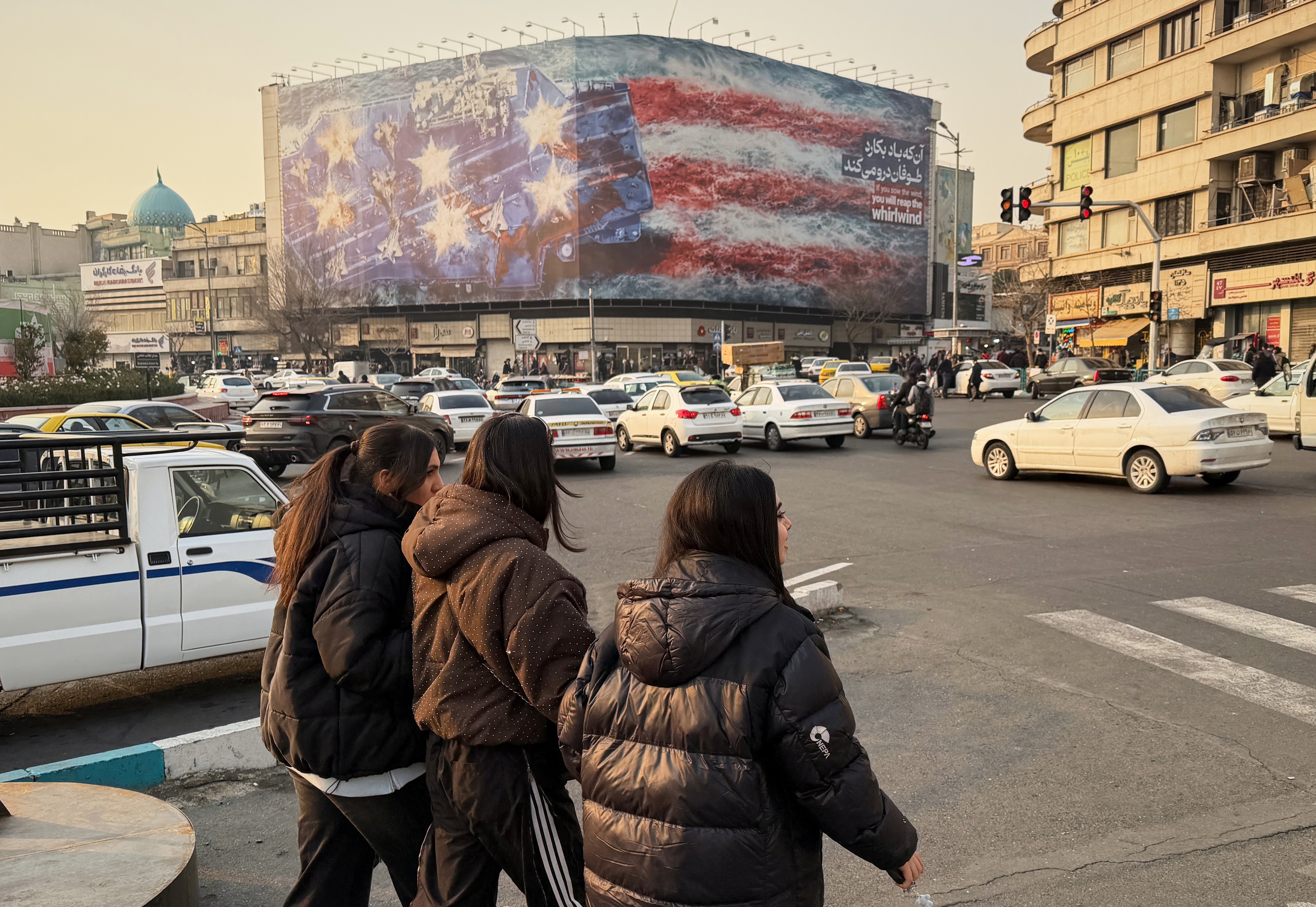 People walk in front a billboard with graphic showing a U.S aircraft carrier with damaged fighter jets on its deck, and sign reading in Farsi and English: "If you sow the wind, you'll reap whirlwind," at the Enqelab-e-Eslami (Islamic Revolution) square, in Tehran, Iran, Sunday, Jan. 25, 2026. (AP)