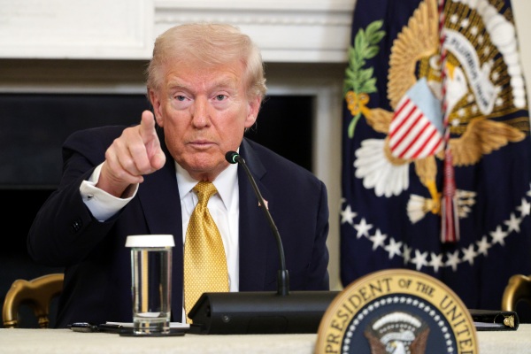 Seated at the White House in front of a microphone, President Trump points with his finger.