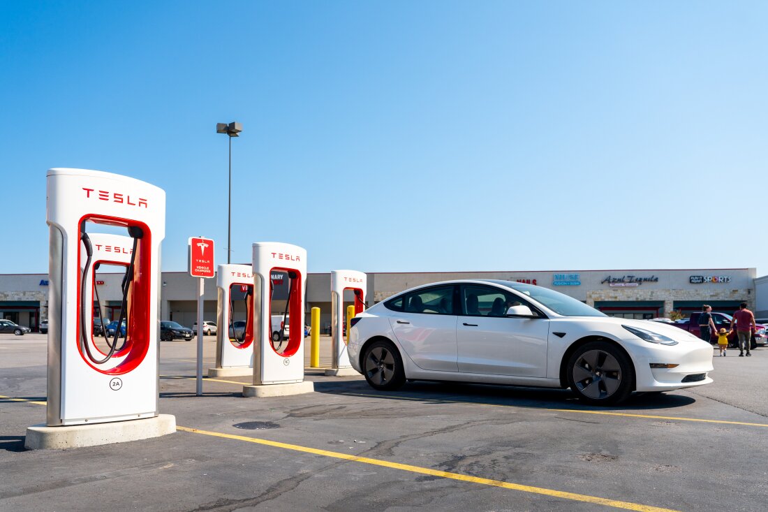 تقف شركة Tesla Superchargers في ساحة انتظار السيارات في أوستن بتكساس في 16 سبتمبر. أربعة من الشواحن الخمسة باللونين الأبيض والأحمر الموجودة في الصورة شاغرة، وتوجد سيارة سيدان بيضاء اللون من طراز تسلا متوقفة أمام إحداها. يمتد مركز تجاري عبر الخلفية.