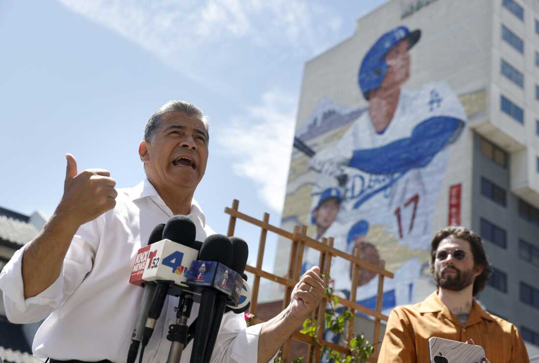 Xavier Becerra is pictured gesturing with news microphones in front of him. In the background, blue sky is visible as well as a huge mural of LA Dodger baseball star Shohei Ohtani.