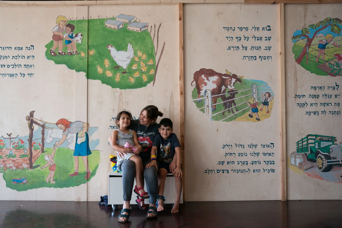 Alice Shahar, 42, poses with her children, Achinoam, 3, and Tzabar, 6, in front of a mural depicting a classic Israeli children's book's pages, originally printed in the Be'eri printing press, the kibbutz's main source of income. Shahar was the coordinator of seven kindergarten classes the displaced kibbutz residents established at the hotel for the Be'eri community. The residents brought the mural with them to stay connected to their home.