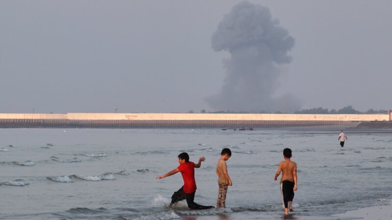 Three boys play in the shallow waters of the Strait of Hormuz, as a plume of smoke rises from an explosion in the background, off Bandar Abbas, Iran, Monday.