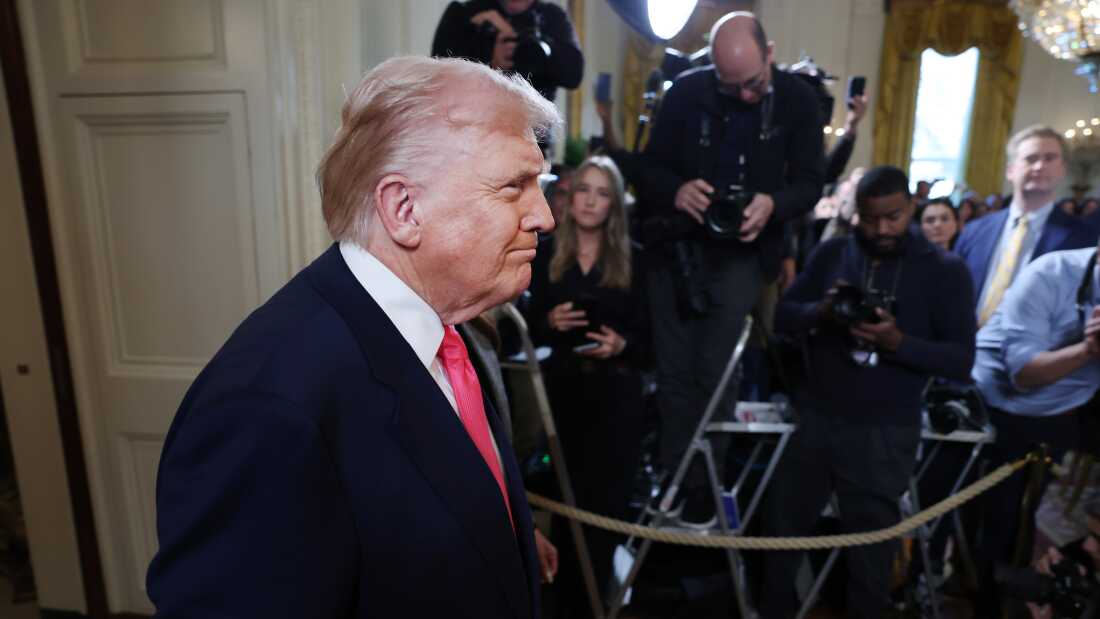 President Trump is seen walking into the East Room wearing a dark blue suit with a red tie. Off to the side, reporters are seen, behind a rope, ready to ask questions and take photos.