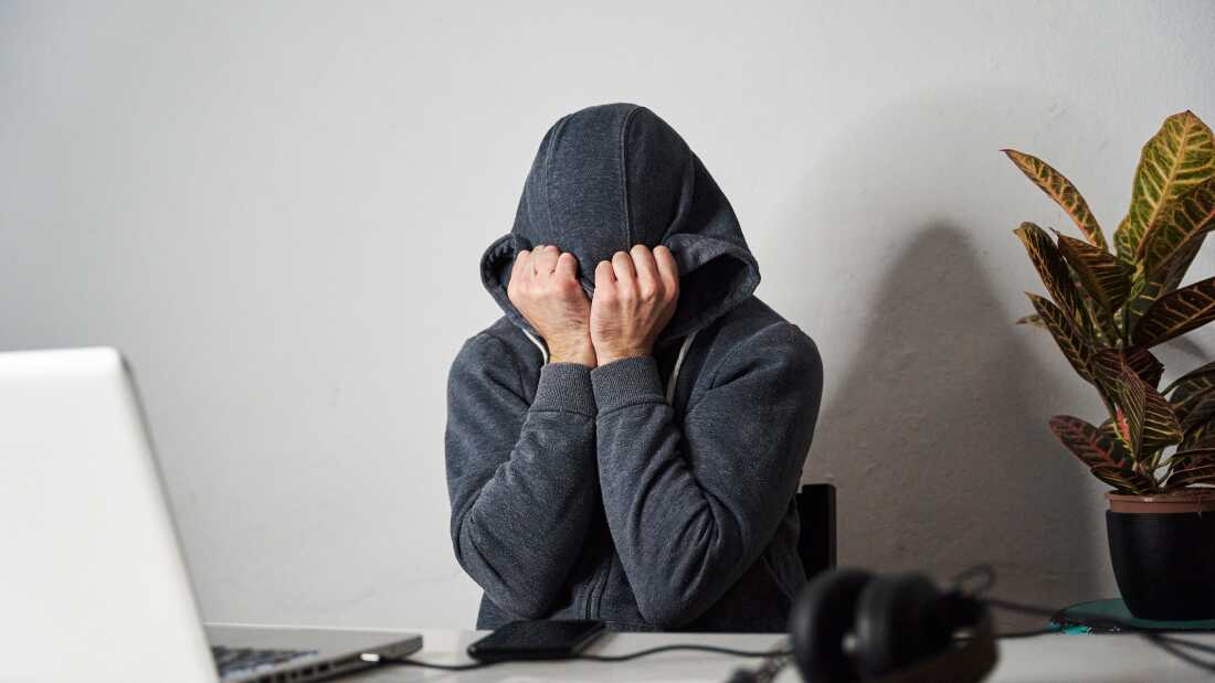 The photo shows a person covering his face with his hands and hoodie at an office desk.