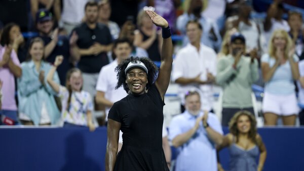 Venus Williams smiles and waves at a crowd. She is wearing an all black tennis dress, black wristbands and a white visor.