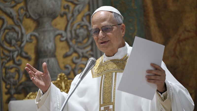 Pope Leo XIV, wearing a white and gold robe, stands in front of a microphone as he concelebrates Mass with the College of Cardinals.