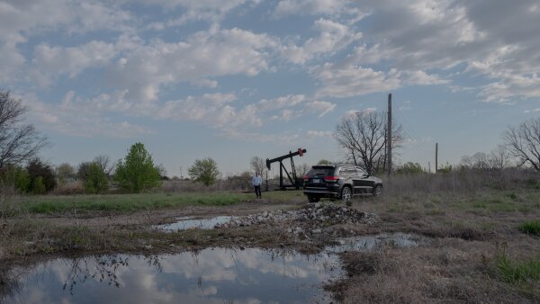 Scott Rabinowitz, president of Grand Resources, stands near a pump jack in Sperry, Okla., on March 20, 2024. Beside the pump is a black SUV. The area is grassy and there is a large puddle of water showing the reflection of the scattered, cloudy sky above. 