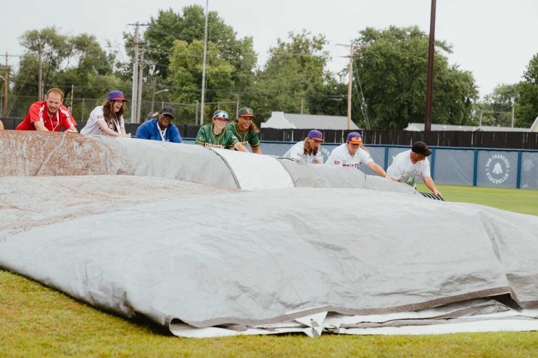 Boston Hunters and San Francisco Firebells players assist the grounds crew in rolling up the infield tarp at Robin Roberts Stadium prior to the start of the inaugural game of the Women's Pro Baseball League. Springfield, IL, August 1, 2026.