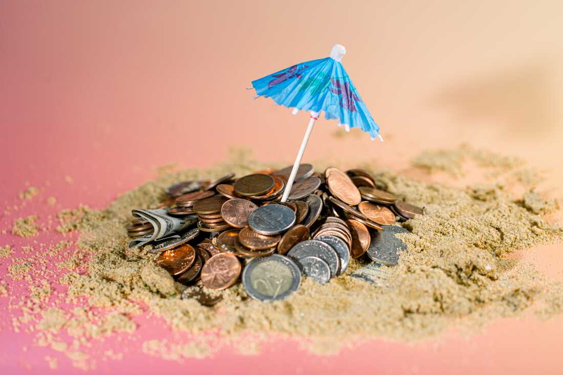 Studio photograph showing a blue paper umbrella propped up in a mound of coins on sand in front of a pink and orange gradient backdrop