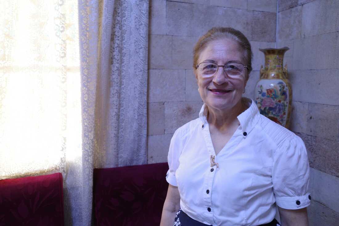 Denise Khoury, age 75, stands in a corner inside the Church of the Sacred Heart of Jesus in Latakia, Syria. Behind her are stone walls, a lace curtain and an urn.