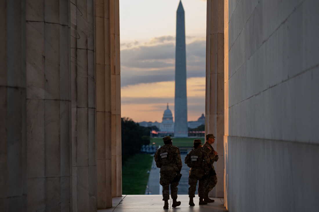 Three National Guard members in uniforms walk along the Lincoln Memorial at sunrise on the National Mall, with the Washington Monument obelisk and the U.S. Capitol dome rising in the distance, in Washington, D.C., on July 23. A row of the Lincoln Memorial's pillars is on the left side of the frame.
