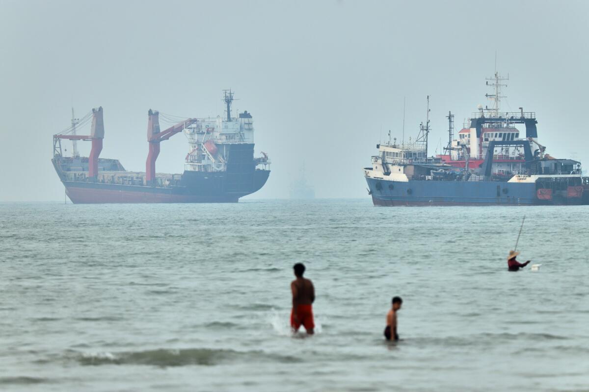 Children wade in the water with cargo ships at anchor in the background and a fisherman nearby, in the Strait of Hormuz off Bandar Abbas, Iran, Tuesday, June 30, 2026.