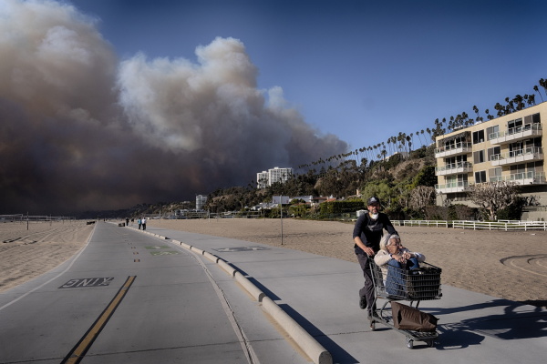A man pushes his mother-in-law in a shopping cart as smoke fills the sky in the distance behind them.