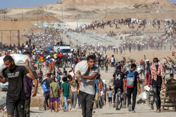 People carry bags of humanitarian aid they received at a distribution center run by the U.S. and Israeli-backed Gaza Humanitarian Foundation (GHF), as they cross the "Netzarim corridor" in the central Gaza Strip, on Aug. 22, while the turrets of Israeli army main battle tanks are pictured in the background.