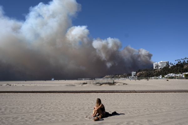A lone sunbather sits on the beach and watches a large plume of smoke from a wildfire rise over the Pacific Palisades.