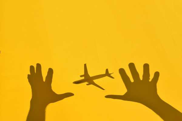 Photograph of a shadow of an airplane and two hards reaching for the airplane, against a yellow wall.
