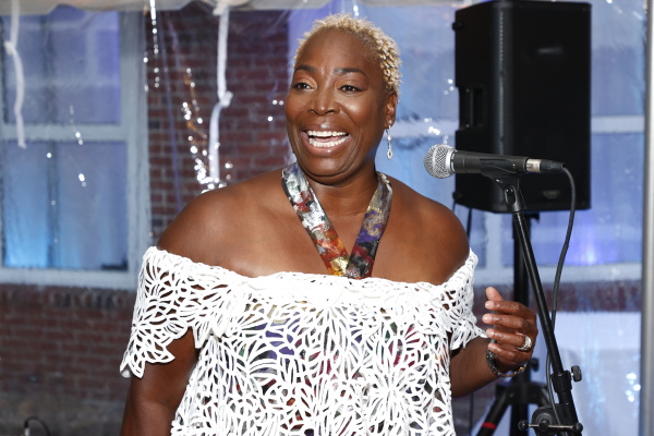 Andrea Lawful-Sanders, wearing a white dress, smiles as she speaks into a microphone.